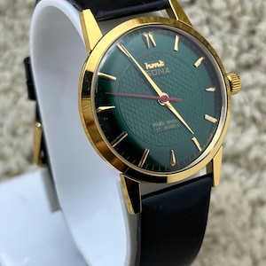 May include: A vintage wristwatch with a gold-tone case and a dark green face. The watch features gold hour markers and hands, a red second hand, and a black leather strap. The watch face has the text "hmt SONA" and "PARA SHOCK 17 JEWELS".