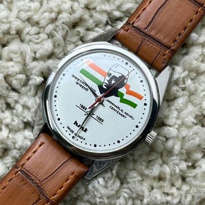 May include: A wristwatch with a white face featuring an image of Jawaharlal Nehru and the Indian flag. The watch has a silver case, black hands, and a brown leather strap. The text "1889-1989" and "Jawaharlal Nehru Centenary" are also visible.
