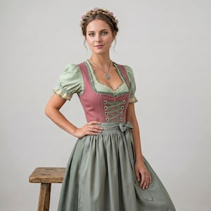 May include: A dirndl dress featuring a dusty rose bodice with a lace-up front, paired with a sage green blouse with puff sleeves and a matching sage green skirt. The outfit includes a floral headpiece and a silver pendant necklace.