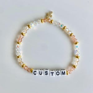 May include: A beaded bracelet with clear, pink, and gold beads. The bracelet has a gold cross charm and white letter beads that spell "CUSTOM". The beads are strung together with gold accents.