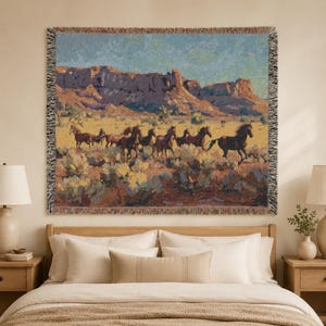 May include: A woven tapestry throw blanket featuring a desert landscape with a herd of horses running across a field of cacti. The scene includes rocky cliffs under a blue sky, finished with a decorative fringed edge on all four sides.