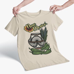 Puede incluir: Camiseta beige con un diseño gráfico colorido. El diseño incluye las palabras "New Mexico" con un girasol, una olla decorativa, maíz y chiles verdes. La camiseta está sujeta por dos manos.