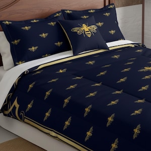 Puede incluir: Juego de cama azul marino con un motivo de abeja dorada. El juego incluye una funda nórdica, fundas de almohada y un cojín con un diseño de abeja grande. La funda nórdica tiene un borde dorado. La ropa de cama está sobre un marco de cama de madera.
