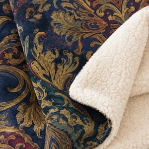 May include: A plush throw blanket featuring a dark blue base with an intricate gold and burgundy damask pattern on one side. The reverse side of the blanket is lined with a thick, cream-coloured, textured sherpa fleece material.