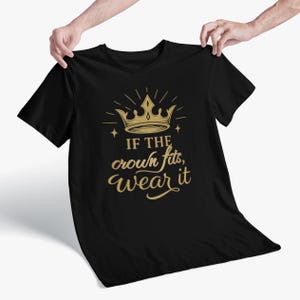 Puede incluir: Camiseta negra con un diseño de corona dorada y la frase "IF THE crown fits, wear it". El diseño incluye una corona, líneas radiantes y estrellas. La camiseta está sujeta por dos manos.