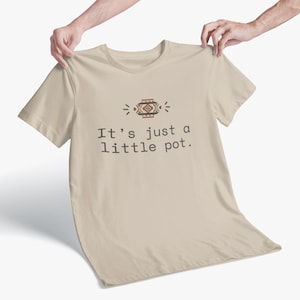 May include: A beige t-shirt with the text "It's just a little pot." The shirt has a decorative graphic above the text with brown, red, and white geometric patterns. The shirt is held up by two hands.