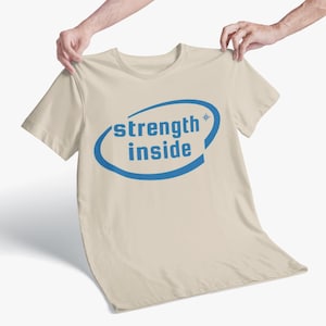 Puede incluir: Una camiseta beige con las palabras "strength inside" en azul, encerradas en un óvalo azul. Un pequeño asterisco está al lado de la palabra "strength". La camiseta está sujeta por dos manos.