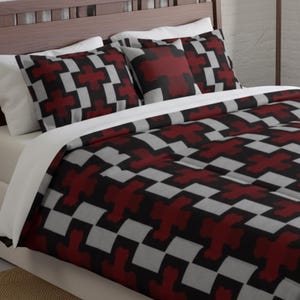 May include: A bed set featuring a duvet cover and pillowcases with a geometric pattern. The pattern consists of red, gray, and black cross-like shapes arranged in a repeating design. The bedding is set on a bed with a wooden headboard.