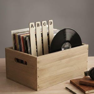 Puede incluir: Una caja de almacenamiento de discos de vinilo de madera llena de discos y divisores etiquetados A, B, C y D. Se ve un disco de vinilo negro. La caja es de madera clara y tiene un recorte de asa en el lateral.