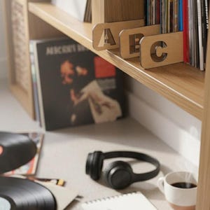 May include: Wooden shelf with vinyl records, dividers labelled "A", "B", and "C", and a pair of black headphones. A cup of coffee and a notepad are also visible. The shelf is made of light-coloured wood.