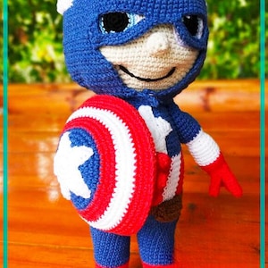 May include: A handmade crochet doll of Captain America, featuring a blue and white mask with the letter "A", a shield, and a red, white, and blue colour scheme. The doll is approximately 25 cm tall.