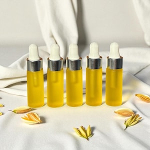 May include: Five small, clear glass dropper bottles filled with a yellow liquid, arranged in a row. Each bottle has a silver cap and a white dropper. Dried yellow flower petals are scattered around the bottles on a white cloth.
