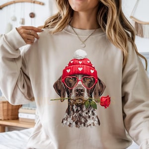May include: Beige sweatshirt with a dog illustration wearing red heart-shaped glasses and a hat with white hearts. The dog holds a red rose. The design is themed for Valentine's Day.