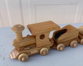 May include: A wooden toy train with a light brown finish. The train features a locomotive, a coal car filled with black pieces, and a caboose. The train has wooden wheels and is designed for children.