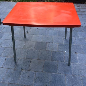 Könnte beinhalten: Ein rechteckiger Tisch mit einer leuchtend orangefarbenen Tischplatte und vier schlanken, geraden Metallbeinen. Die Oberfläche ist glatt und flach und weist abgerundete Ecken auf. Die Beine sind an jeder Ecke angebracht.