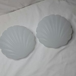 May include: Two frosted glass seashell-shaped light covers. The covers are white and have a scalloped edge with a ribbed design. The light covers are on a white background.