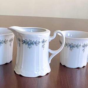 May include: A white porcelain creamer and two matching cups. Each piece features a delicate blue floral design around the rim. The creamer has a handle and spout, while the cups have curved handles. The set is displayed on a wooden surface.