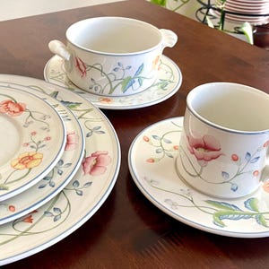 May include: A white porcelain dinner service with a floral pattern. The set includes plates, a soup bowl, and a teacup, all decorated with pink, yellow, and green flowers and a blue trim. The plates are of varying sizes, suitable for a meal.