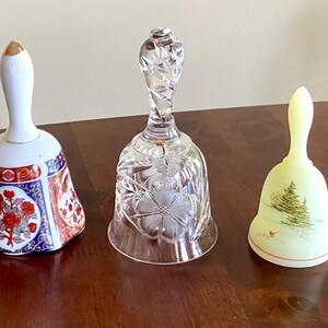 May include: Three decorative bells on a wooden surface. The bells include a porcelain bell with floral designs, a clear crystal bell, and a pale yellow bell with a painted Christmas tree design.