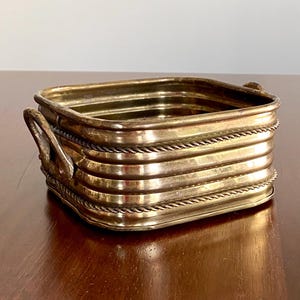 May include: A square brass container with rounded corners and two side handles. The exterior features horizontal ribbed detailing with a rope-like texture between the ridges. The metal has a polished gold-toned finish with visible reflections.