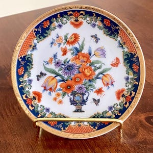 May include: Decorative porcelain plate with a floral arrangement in a vase, featuring orange, purple, and blue flowers, and butterflies. The plate has a gold rim and is displayed on a gold stand. The plate's border is orange and blue.