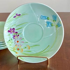 May include: A set of light green ceramic saucers with floral designs. Each saucer features a central indentation and is decorated with hand-painted flowers in shades of purple, orange, and blue. The saucers are displayed on a brown surface.