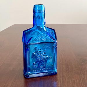May include: A cobalt blue glass bottle with a rectangular body and a tall, square neck. The front of the bottle features a raised relief of a man on horseback, with the text "1775" and "PAUL REVERE".