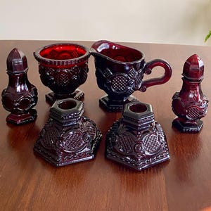 May include: A collection of six deep red pressed glass items including a creamer with a handle, a sugar bowl, two salt and pepper shakers, and two candle holders. Each piece features a repeating geometric pattern with circular and diamond motifs.