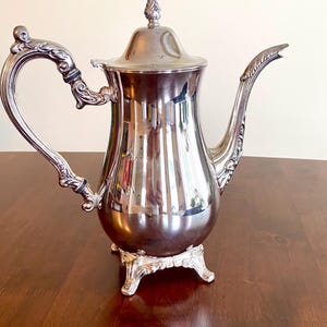 May include: A silver-colored coffee pot with a lid and ornate handle and spout. The pot sits on a decorative base with three feet. The pot is highly reflective, showing the surrounding environment. The handle has a black accent.
