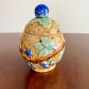 May include: A ceramic jar with a lid, featuring a basket weave design in tan with green leaves and blue berries. The lid is topped with a blue berry-shaped knob. The jar is sitting on a brown surface.