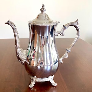 May include: A polished silver coffee pot with a decorative handle and spout. The pot has a lid with a small finial and sits on three ornate feet. The reflective surface of the pot mirrors the surrounding environment, creating a vintage aesthetic.