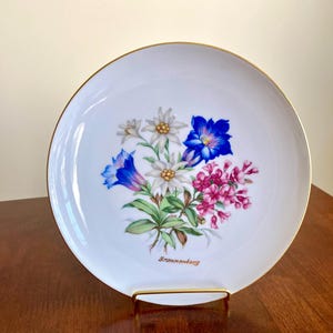 May include: White porcelain plate with a gold rim, featuring a floral bouquet of blue, white, and pink flowers. The word "Brannenburg" is written below the flowers. The plate is displayed on a gold-colored stand.