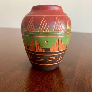 May include: A small, handcrafted ceramic vase with a rounded body and a narrow neck. The vase features a color scheme of reddish-brown, green, orange, and black. The design includes carved lines and geometric patterns, adding a touch of rustic charm to the decorative piece.