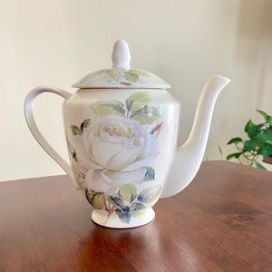 Könnte beinhalten: Cremefarbene Keramik-Teekanne mit weißem Rosenblumendesign. Die Teekanne hat einen gebogenen Griff, einen langen Ausguss und einen Deckel mit einem kleinen Knopf. Das Rosendesign umfasst grüne Blätter und Stiele.