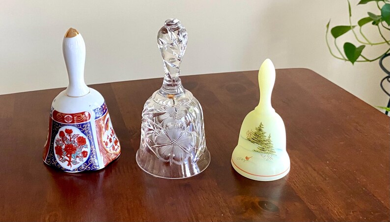 May include: Three decorative bells on a wooden surface. The bells include a porcelain bell with floral designs, a clear crystal bell, and a pale yellow bell with a painted Christmas tree design.