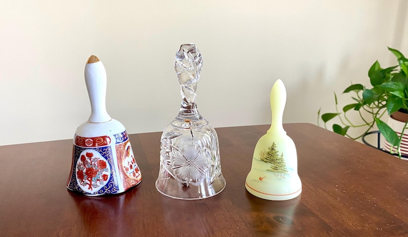 May include: Three decorative bells on a wooden surface. The bells include a porcelain bell with a floral design, a clear crystal bell, and a pale yellow bell with a Christmas tree design. The bells are of varying sizes and shapes.