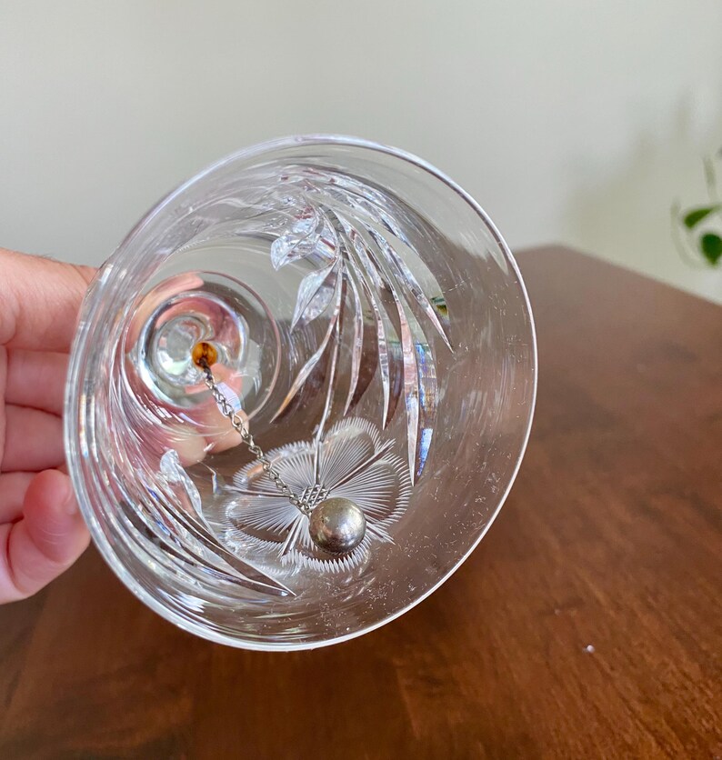 May include: Clear glass bell with etched floral and leaf designs. A small silver ball hangs inside the bell, suspended by a delicate chain. A small amber-colored bead is at the top of the chain.