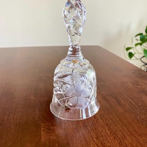 May include: A clear glass bell with a floral design etched on the body and handle. The handle has a twisted design. The bell is sitting on a brown wooden surface.