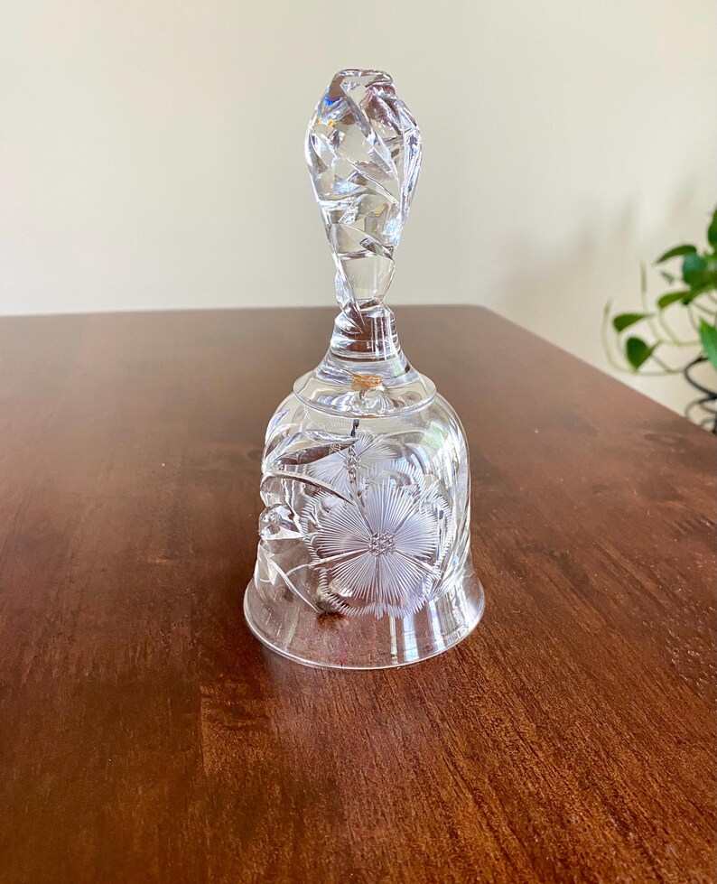 May include: A clear glass bell with a floral design etched on the body and handle. The handle has a twisted design. The bell is sitting on a brown wooden surface.