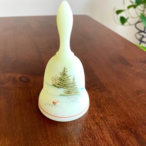 May include: A pale green bell with a long handle, featuring a winter scene. The bell has a painted image of a Christmas tree, snow, and a red bird. A thin red line accents the bottom edge. The bell is likely made of glass or ceramic.