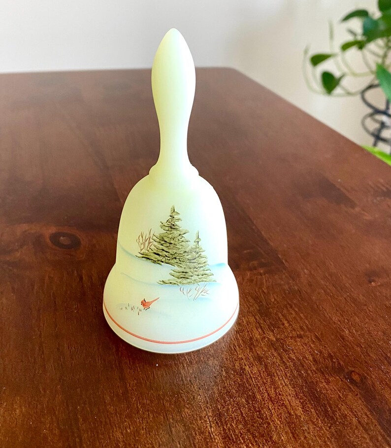 May include: A pale green bell with a long handle, featuring a winter scene. The bell has a painted image of a Christmas tree, snow, and a red bird. A thin red line accents the bottom edge. The bell is likely made of glass or ceramic.