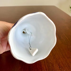 May include: A white ceramic bell with a scalloped edge. The bell's exterior features a design with red and blue accents. A small, triangular clapper hangs inside the bell. The bell is held in a person's hand.
