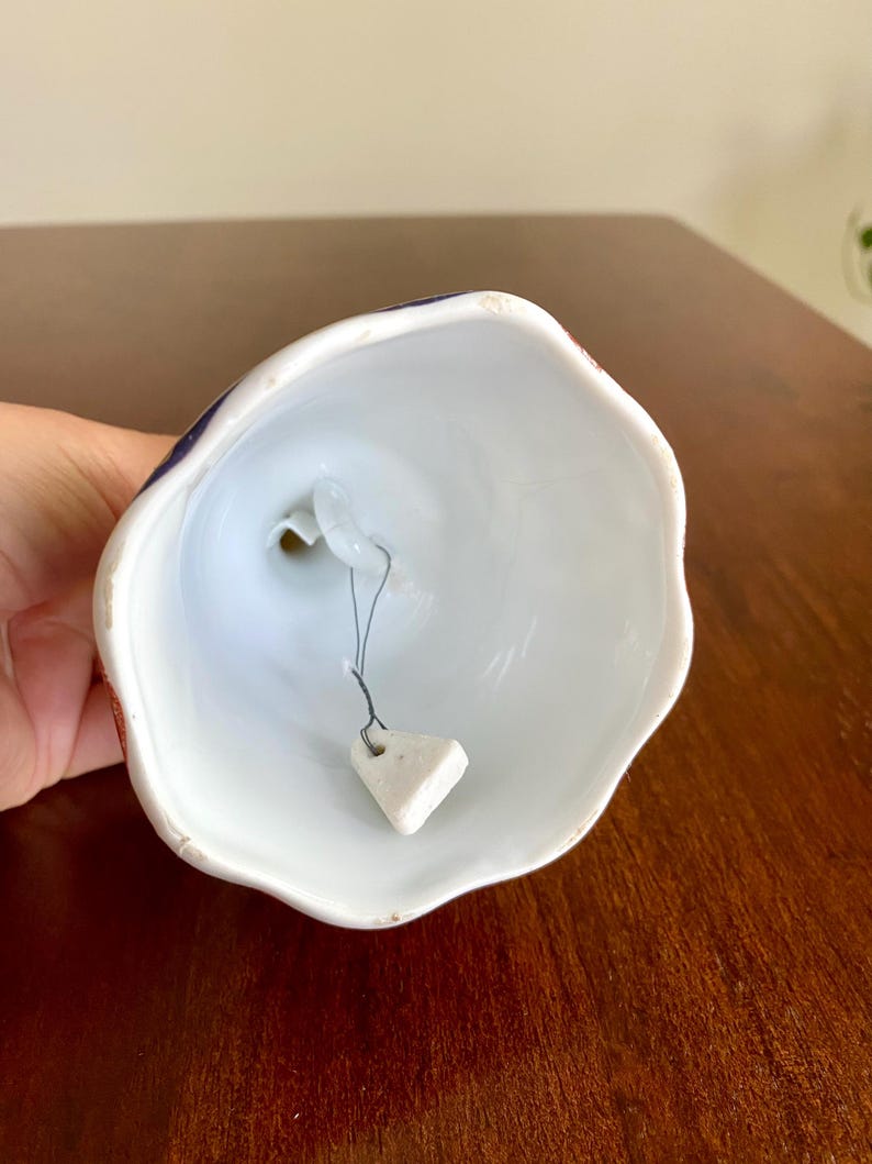 May include: A white ceramic bell with a scalloped edge. The bell's exterior features a design with red and blue accents. A small, triangular clapper hangs inside the bell. The bell is held in a person's hand.