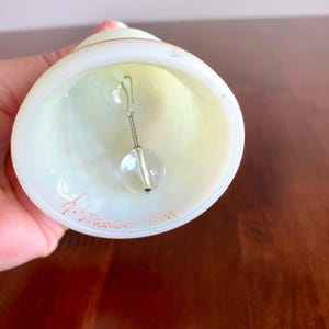 May include: A hand-blown glass bell with a clear, spherical clapper suspended inside. The bell is white with a pale yellow interior and a red accent near the top. The bell has the signature "J. Stevens" and "ICM" etched on the bottom.