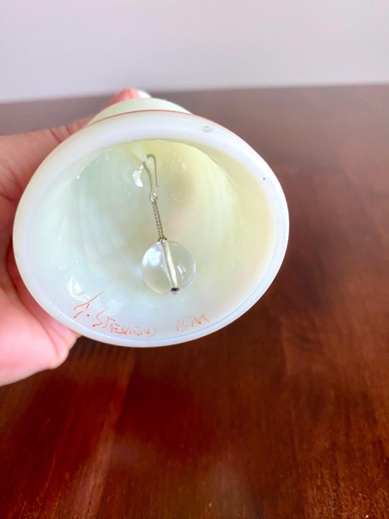 May include: A hand-blown glass bell with a clear, spherical clapper suspended inside. The bell is white with a pale yellow interior and a red accent near the top. The bell has the signature "J. Stevens" and "ICM" etched on the bottom.