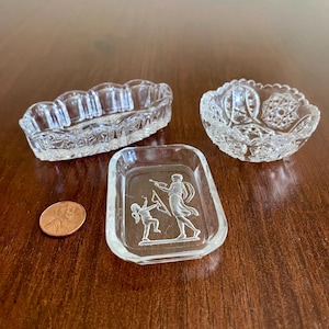 May include: Three clear glass dishes of varying shapes and a copper penny. One rectangular dish features an etched design of two figures, one holding a bow and arrow. Another is an oval scalloped dish, and the third is a round bowl with a star pattern.