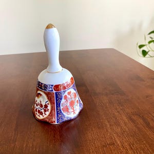 May include: A white ceramic bell with a gold-tipped handle and a flared base. The base features red, blue, and gold floral designs. The bell is likely a decorative item.