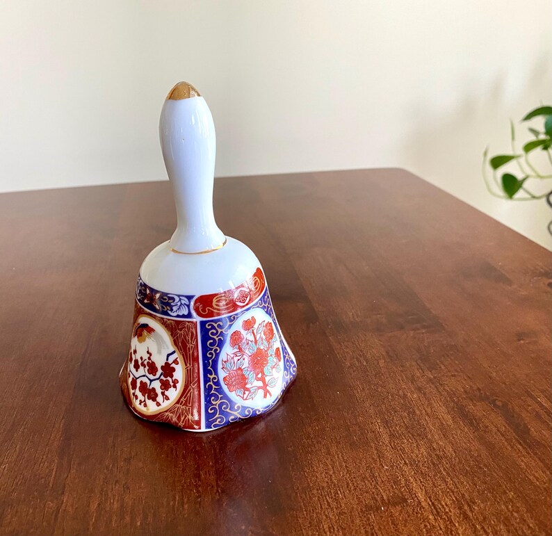 May include: A white ceramic bell with a gold-tipped handle and a flared base. The base features red, blue, and gold floral designs. The bell is likely a decorative item.