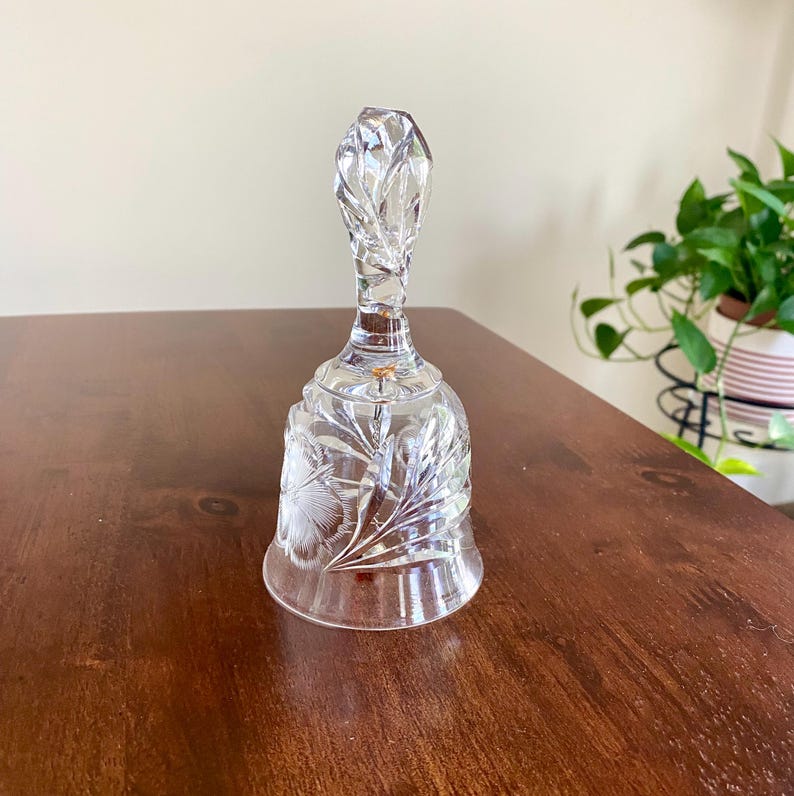 May include: A clear glass bell with a faceted handle and floral etched design. The bell is sitting on a dark brown wooden surface. The bell is a decorative item.