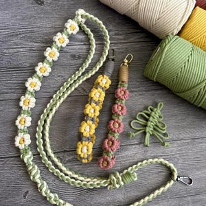 Puede incluir: Lanyard y llaveros de macramé con margaritas blancas, amarillas y rosas. El lanyard es verde y blanco con detalles de margaritas. Los llaveros presentan diseños de margaritas amarillas y rosas. También hay un pequeño árbol de macramé verde.